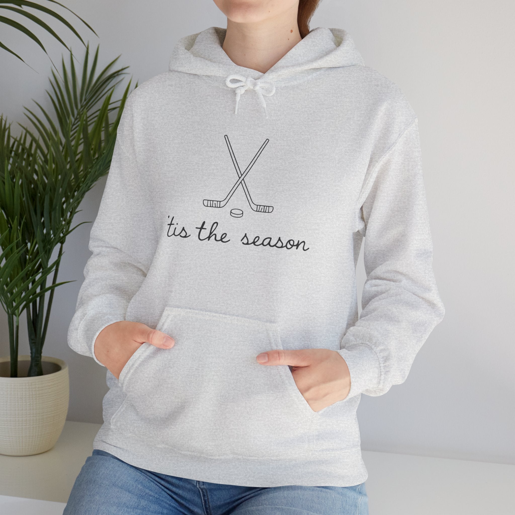 Person wearing a gray hoodie with 'tis the season and hockey stick design, standing against a plain background.