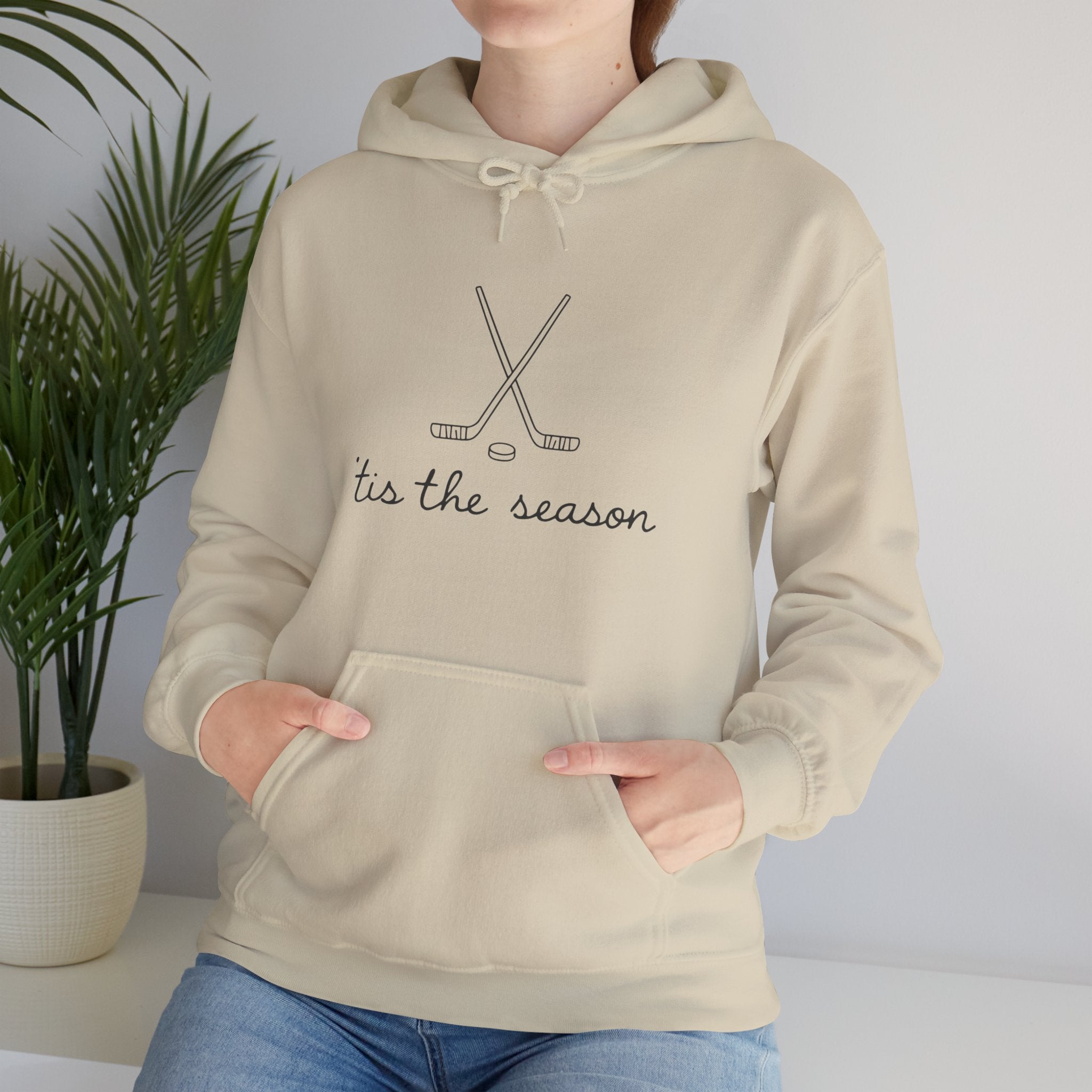 Beige hoodie with hockey design and text 'tis the season worn by a person, with a plant in the background.