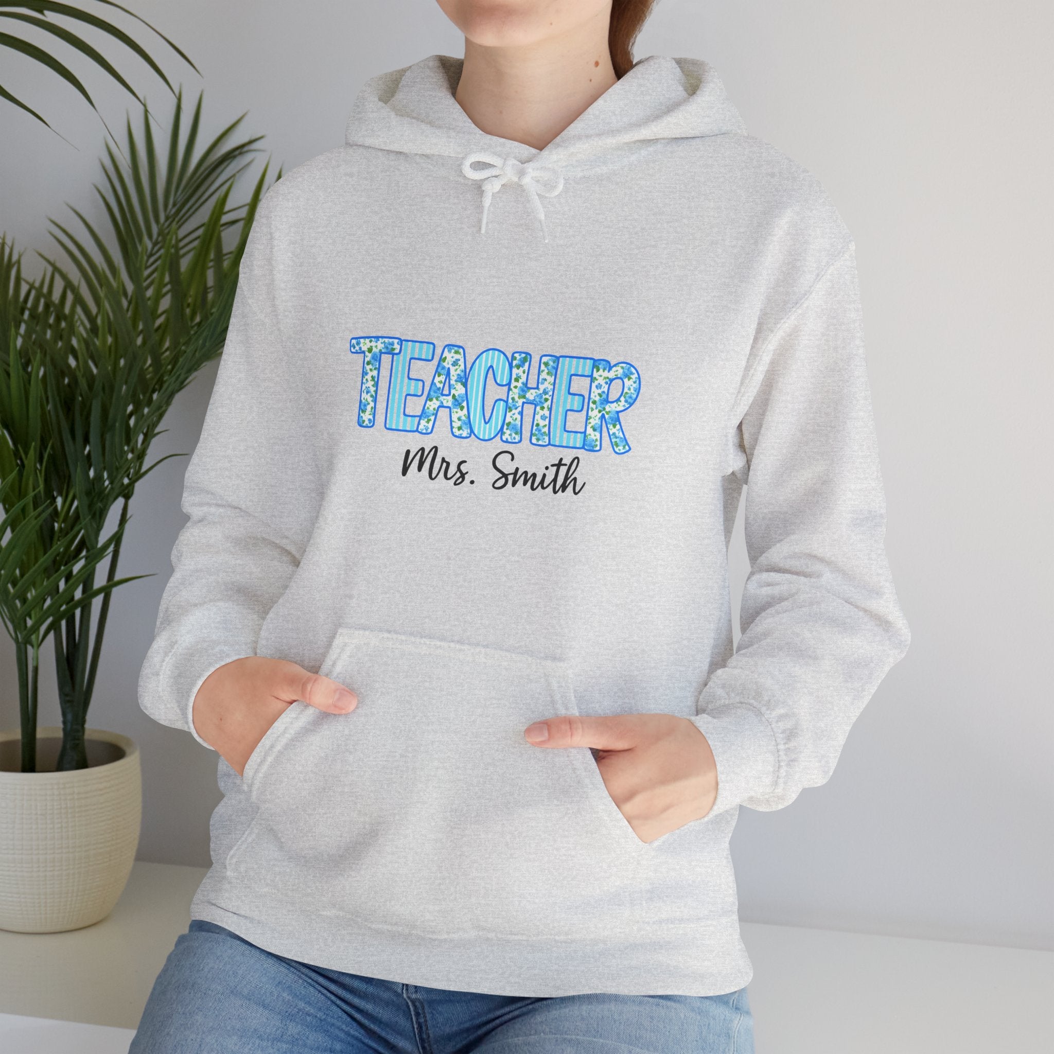 Person wearing a gray hoodie with 'TEACHER Mrs. Smith' text, standing indoors with a plant in the background.